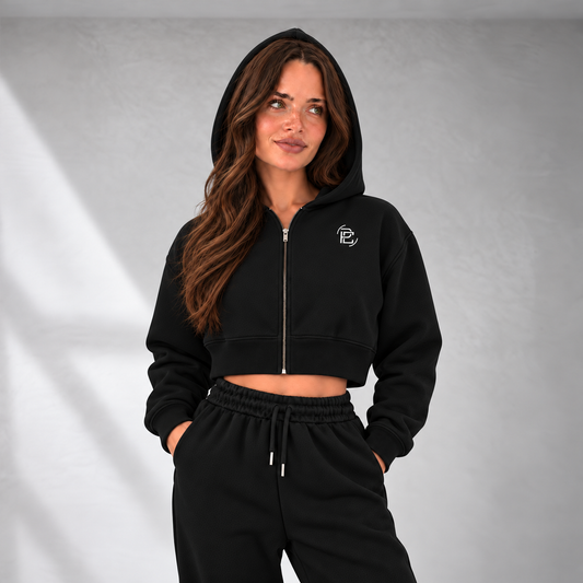 A woman wearing a black cropped zip-up hoodie with a small white embroidered logo on the left chest, paired with matching black sweatpants, standing against a gray textured wall.