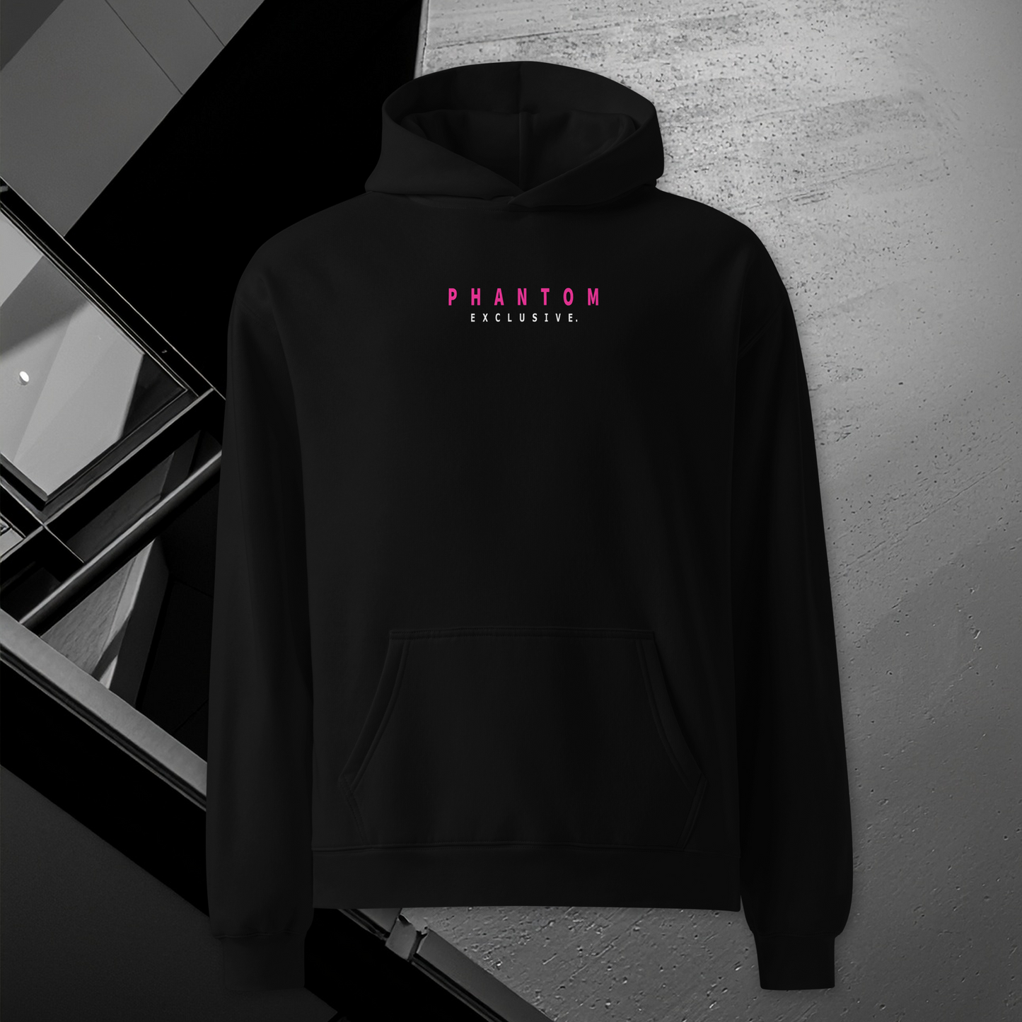 A black pullover hoodie with 'PHANTOM EXCLUSIVE' printed in pink and white on the chest, displayed against a textured gray concrete wall with a modern black window frame.