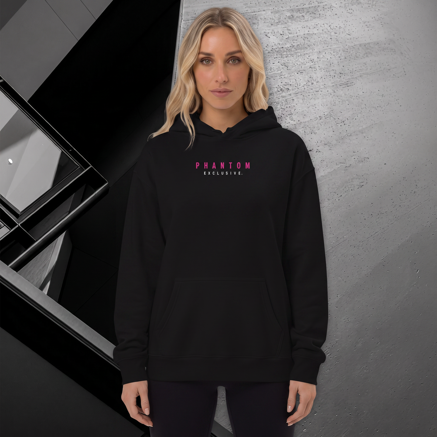 A blonde woman wearing a black pullover hoodie with 'PHANTOM EXCLUSIVE.' printed in pink and white on the chest, standing against a textured gray wall.
