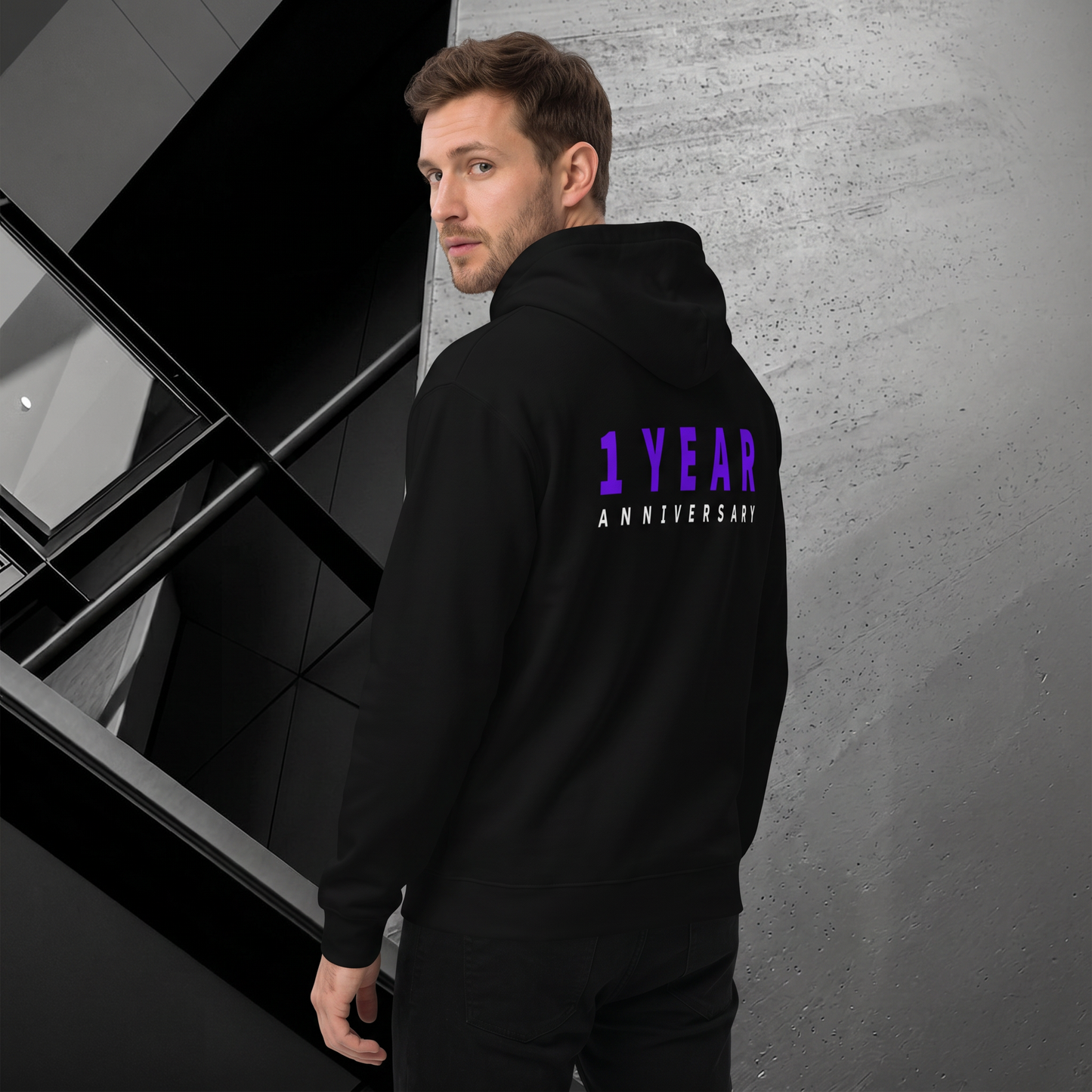 A man wearing a black hoodie with '1 YEAR ANNIVERSARY' printed in purple and white on the back, standing in a modern interior setting.