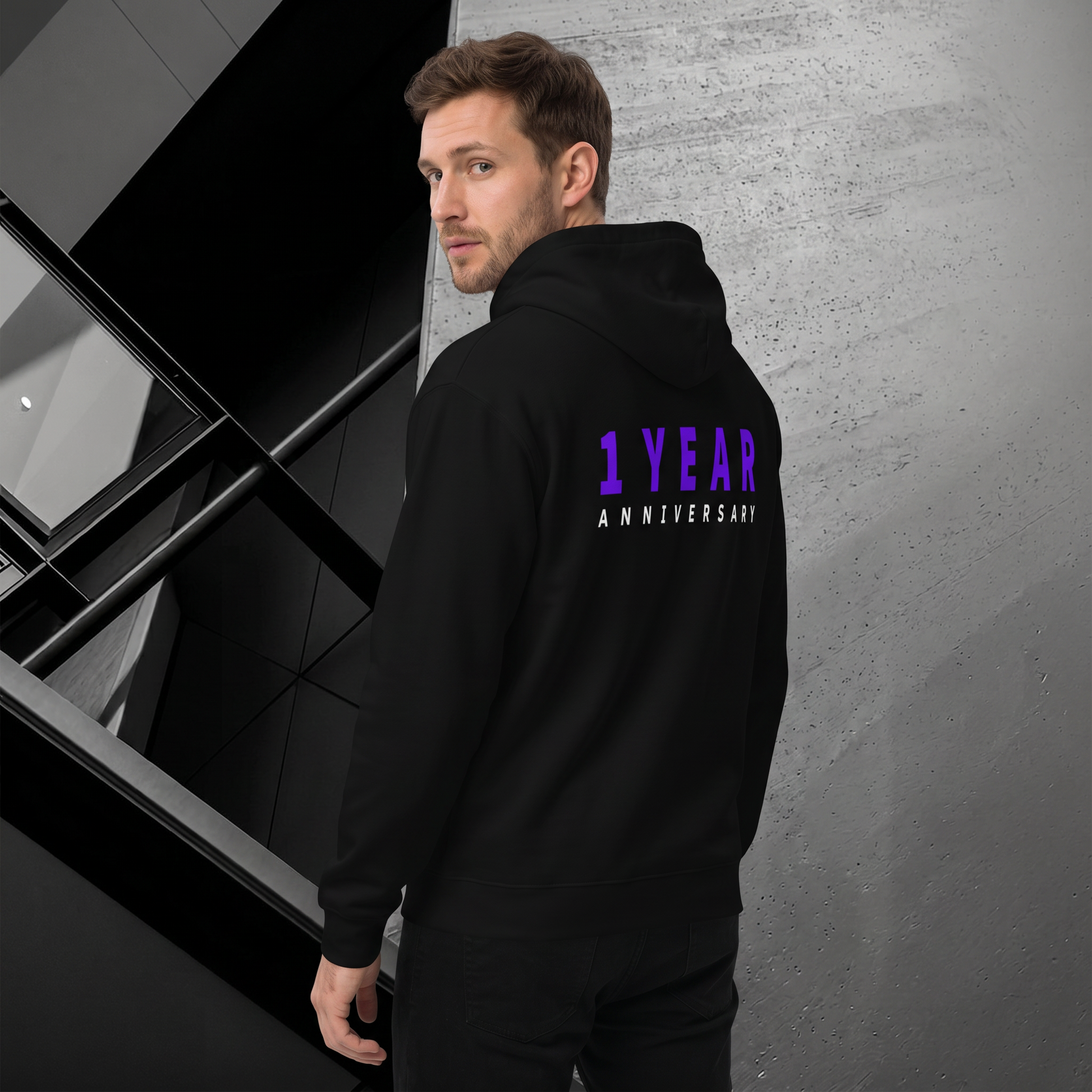 A man wearing a black hoodie with '1 YEAR ANNIVERSARY' printed in purple and white on the back, standing in a modern interior setting.