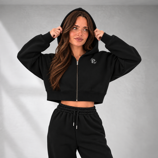 A woman wearing a black cropped zip-up hoodie with a small white embroidered logo on the chest, paired with black sweatpants, posing against a gray studio background.