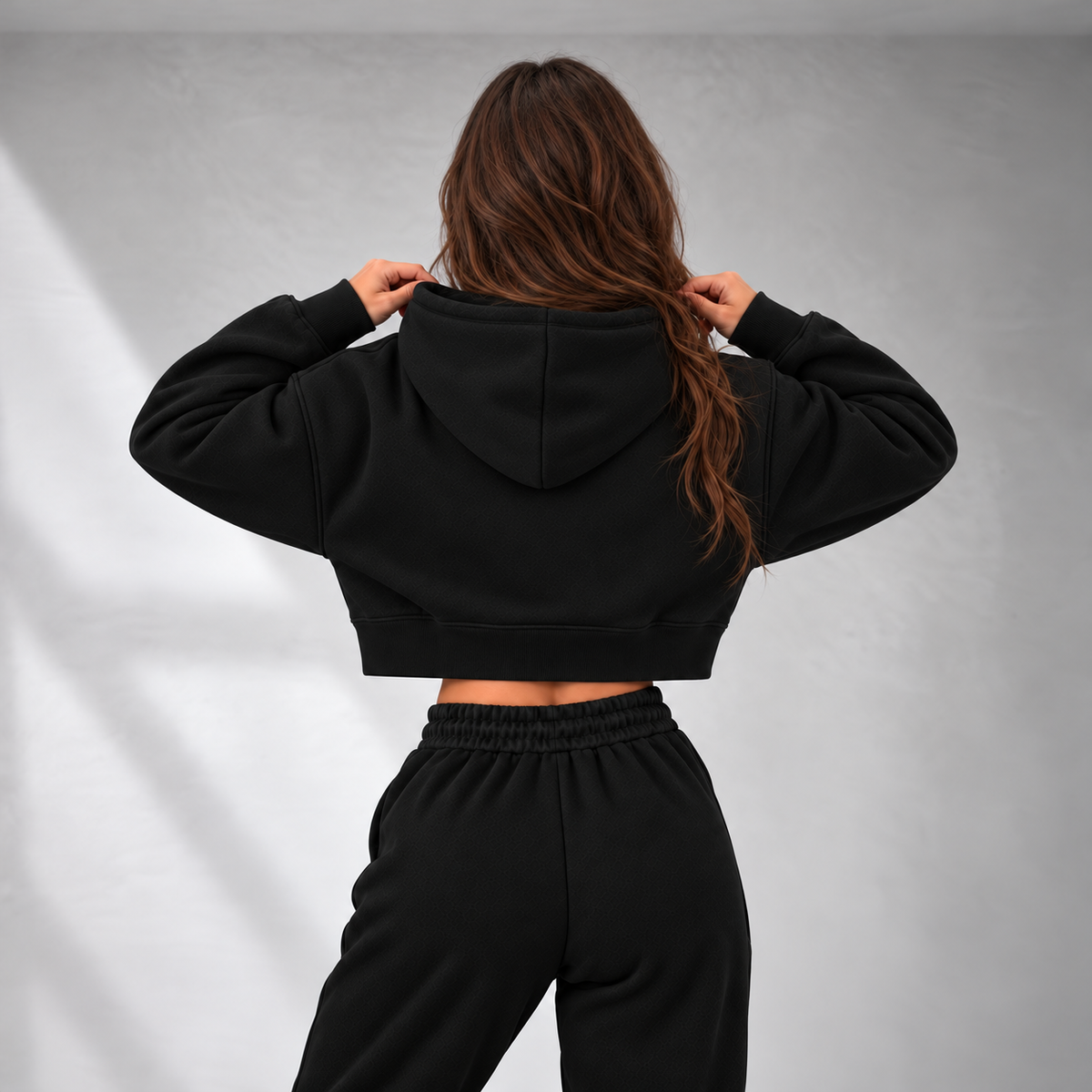 A woman viewed from behind wearing a black cropped hoodie and matching black sweatpants against a light gray studio background.