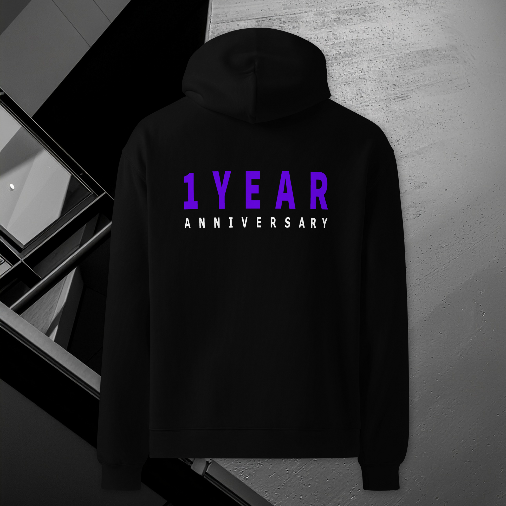 A black pullover hoodie shown from the back, featuring the text '1 YEAR ANNIVERSARY' printed on the back in purple and white. The hoodie is displayed against a dark, modern architectural background.