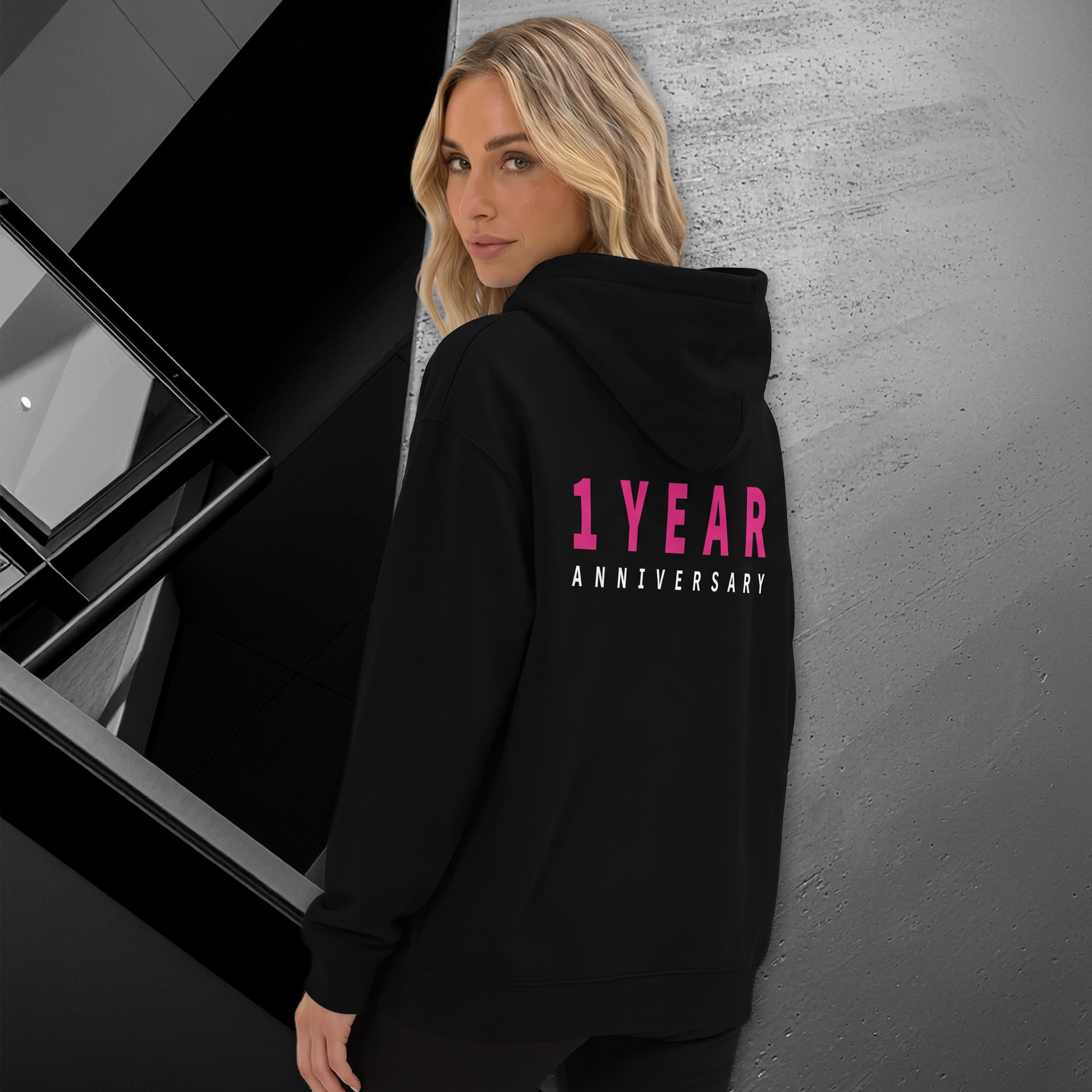 A woman wearing a black hoodie with '1 YEAR ANNIVERSARY' printed in pink and white on the back, leaning against a textured gray wall in a modern interior setting.