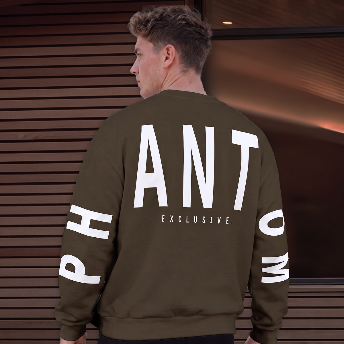 A man viewed from behind wearing an olive brown crewneck sweatshirt with large white text on the back reading 'ANT' and 'EXCLUSIVE.' plus partial sleeve text, set against a dark wood slat wall.